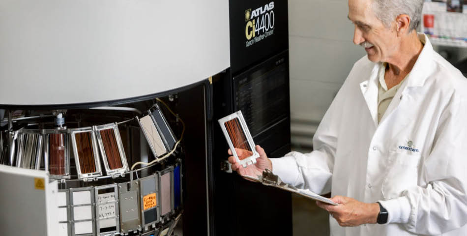 A man in a lab coat examines a rectangular sample in front of a large testing machine, holding a clipboard and smiling.