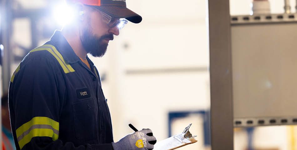 A worker wearing safety gloves, glasses, and a hard hat writes on a clipboard in an industrial setting with bright lighting.