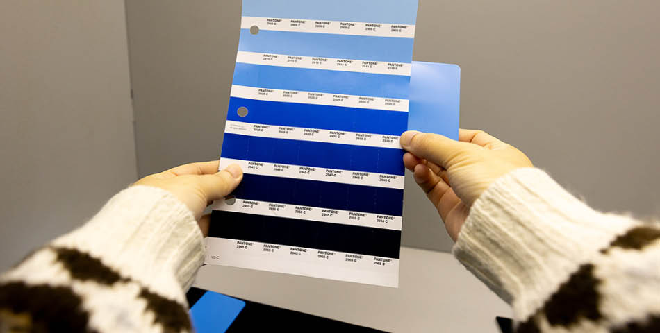A person holds a color swatch card displaying multiple shades of blue, each labeled with codes, against a plain background.