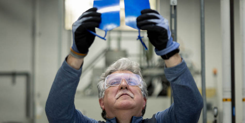 An older man wearing safety goggles and gloves inspects two blue plastic panels, holding them up to the light in an industrial setting.