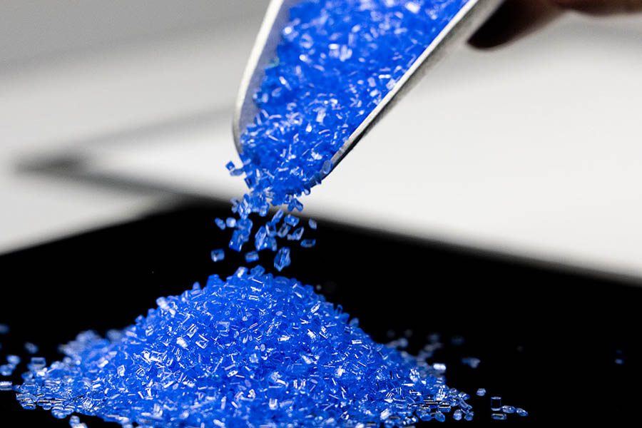 A metal scoop pours a pile of small blue plastic pellets onto a flat black surface.