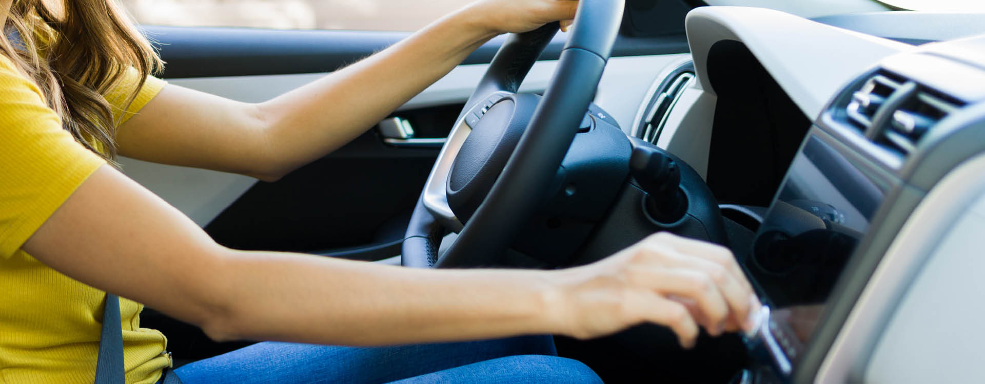 Person in a yellow shirt sitting in the driver’s seat of a car, one hand on the steering wheel and the other adjusting the car’s dashboard controls.