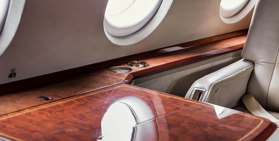 Close-up view of a polished wooden table and leather seat inside a private jet, with round windows visible along the wall.