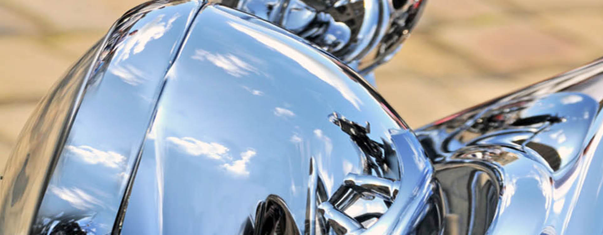 Close-up of shiny chrome parts of a motorcycle reflecting clouds and blue sky, with a blurred cobblestone ground in the background.