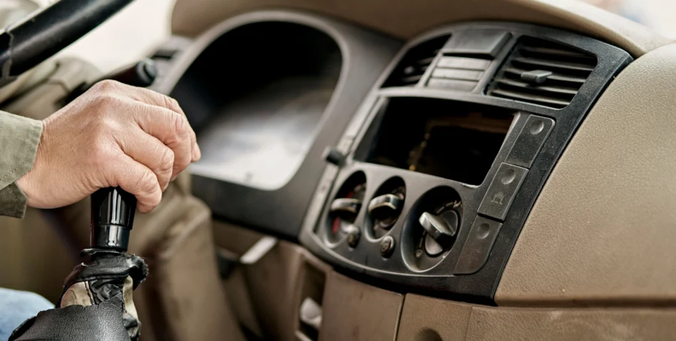 A person’s hand shifts the gear lever inside a vehicle, with the dashboard, air vents, and climate control knobs visible.