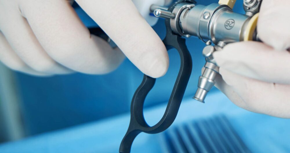 Close-up of a gloved hand holding a medical endoscope, with surgical instruments visible in the background.
