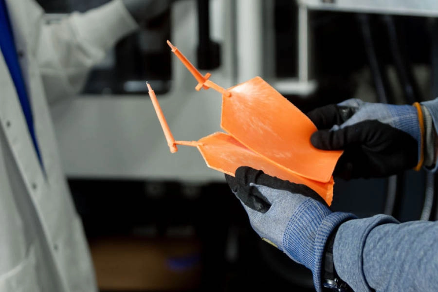 A person wearing gloves holds two orange plastic molded parts with sprues, while another person in a lab coat stands nearby.