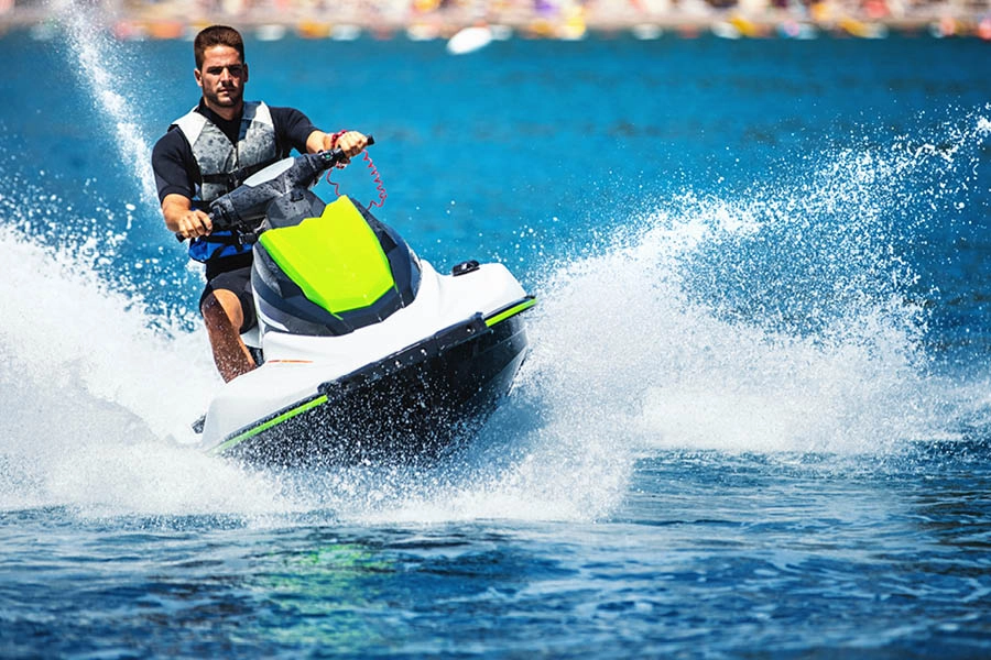 A man wearing a life jacket rides a green and white jet ski on blue water, creating splashes as he speeds forward.