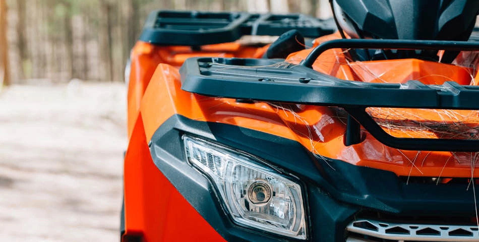 Close-up of the front section of an orange ATV, showing the headlight, front rack, and part of the hood, with a blurred outdoor background.