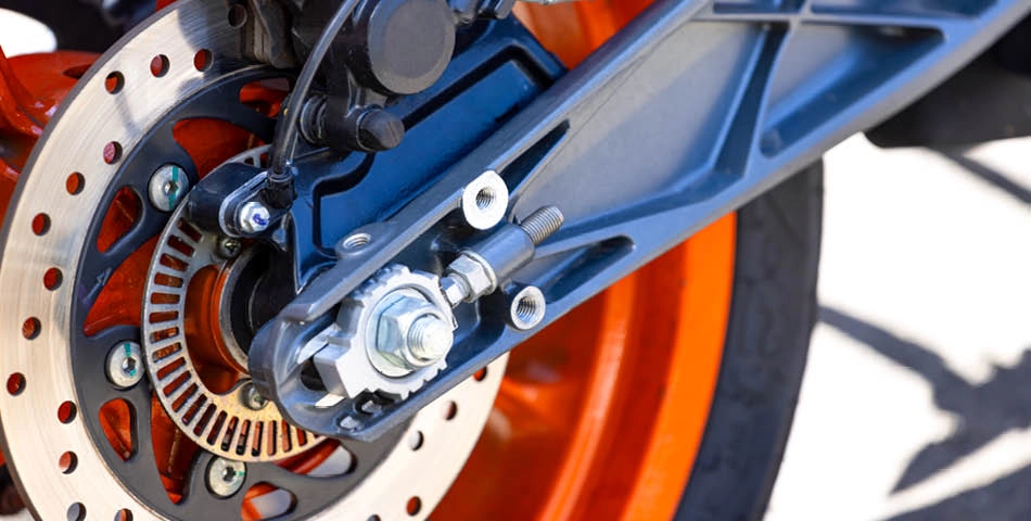 Close-up of a motorcycle rear wheel showing the disc brake, caliper, axle, and spokes, with orange rim and metallic components clearly visible.