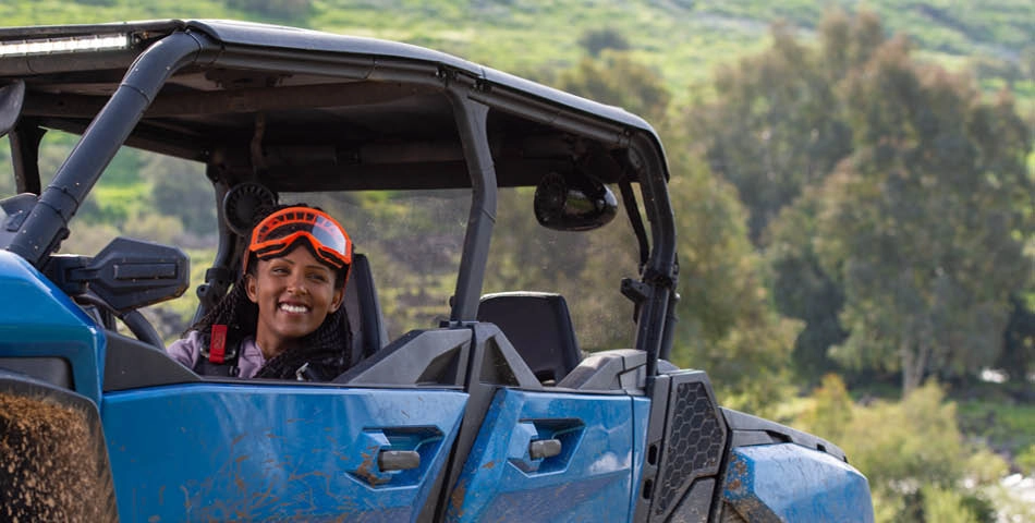 A person wearing orange goggles sits and smiles in the driver's seat of a blue off-road vehicle, with greenery visible in the background.