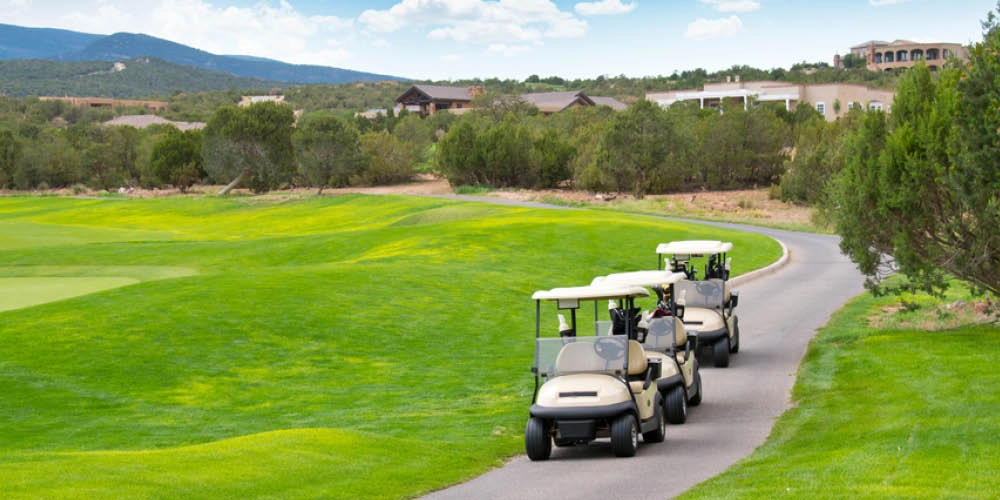 Three golf carts drive along a paved path on a golf course surrounded by green grass, trees, and distant buildings under a partly cloudy sky.