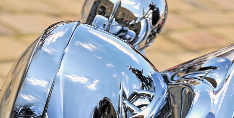 Close-up of shiny chrome motorcycle parts reflecting clouds, sky, and nearby surroundings on a cobblestone surface.