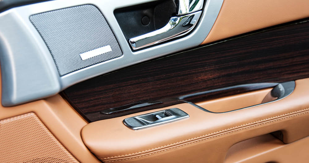 Close-up of a car’s interior door panel featuring tan leather, dark wood trim, and silver accents, including window and lock controls.