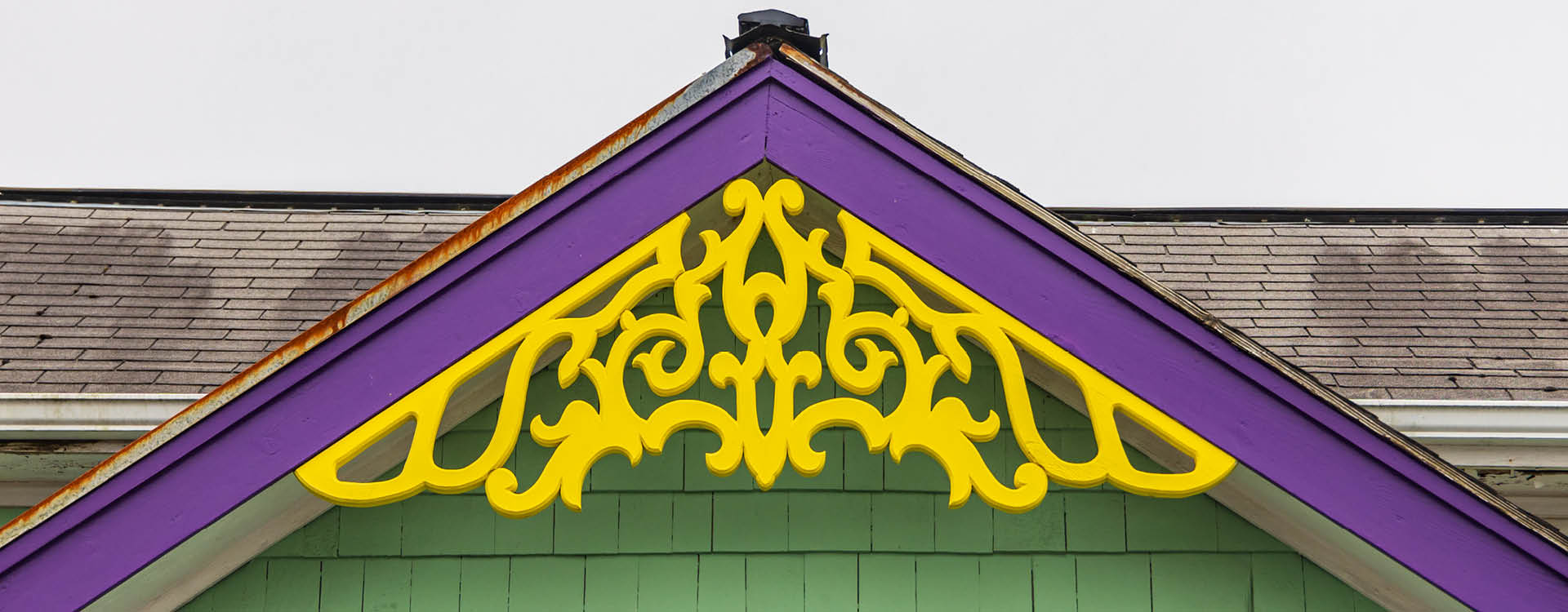 A house gable with decorative yellow trim, purple eaves, green siding, and gray shingles on the roof.