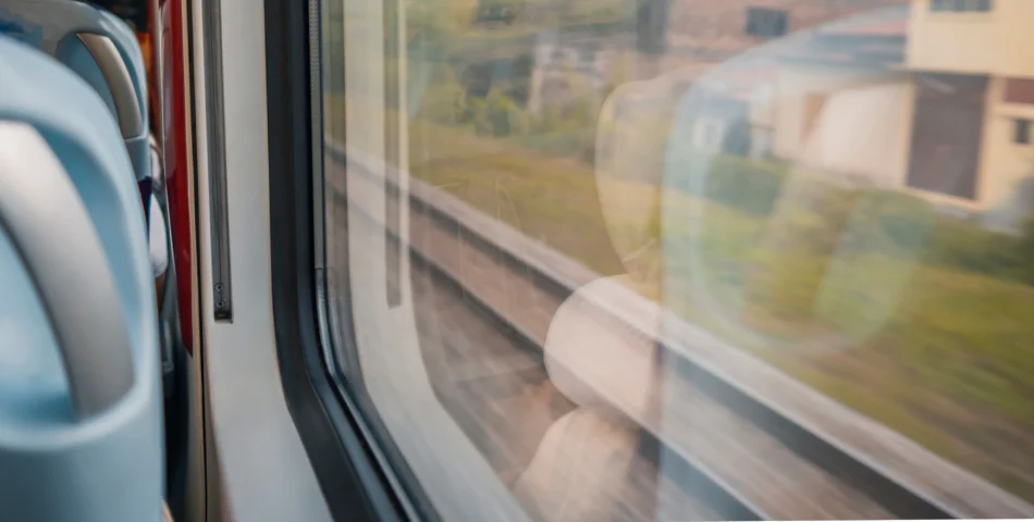 View from a train window showing blurred railway tracks and green landscape outside, with reflections of seats visible on the glass.