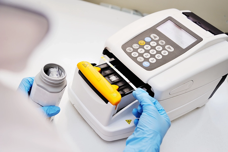 A person wearing blue gloves operates a laboratory machine, placing a sample container inside its slot for analysis.