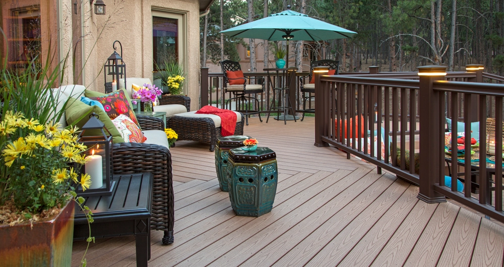 Spacious outdoor deck with wicker furniture, colorful cushions, potted plants, decorative stools, and a dining set under a large teal umbrella, surrounded by a brown railing.
