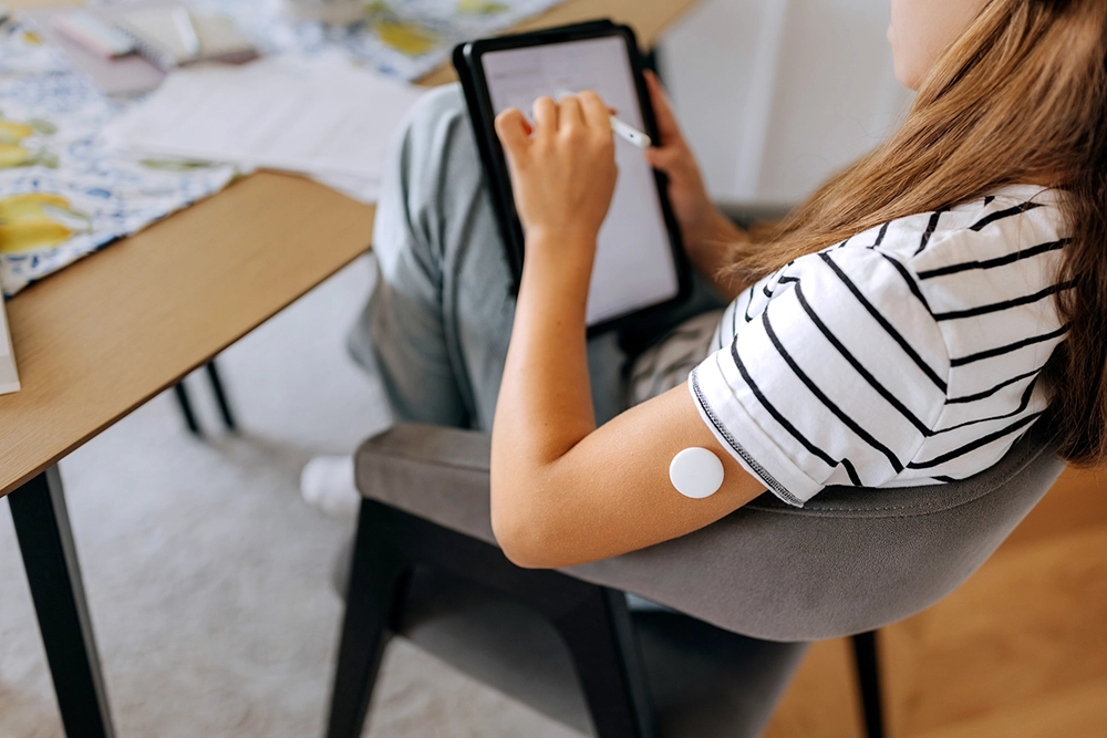 A person in a striped shirt sits at a table using a tablet, with a circular sensor attached to their upper arm.