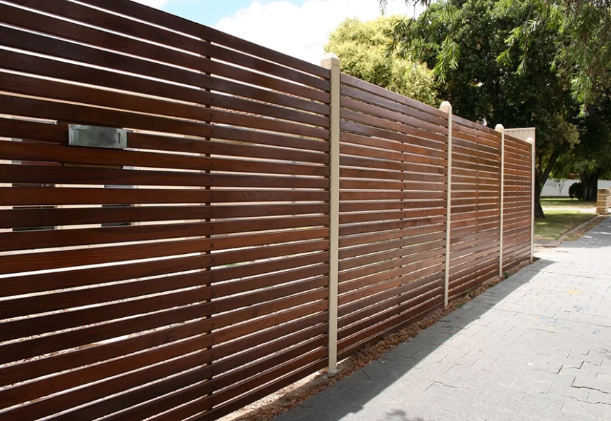A modern wooden slat fence with vertical beige posts stands along a paved sidewalk beside trees and grass.