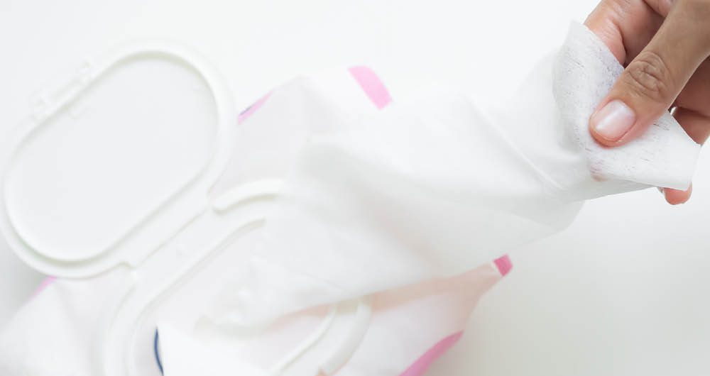 A hand pulling a wet wipe from an open plastic container of wipes on a white surface.