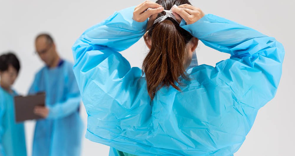 Person in blue protective gown ties hair covering; two other individuals in similar attire are in the background, one holding a clipboard.