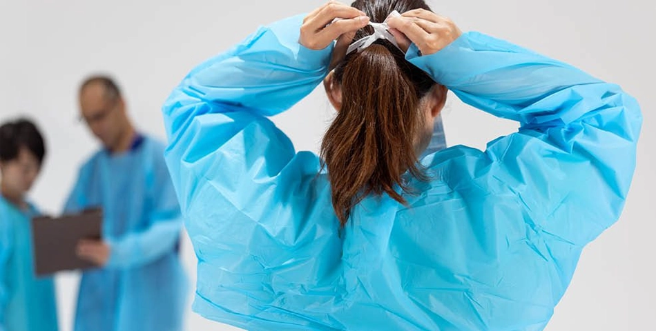 A person in blue protective clothing ties the back of their gown, with two others in similar attire blurred in the background.