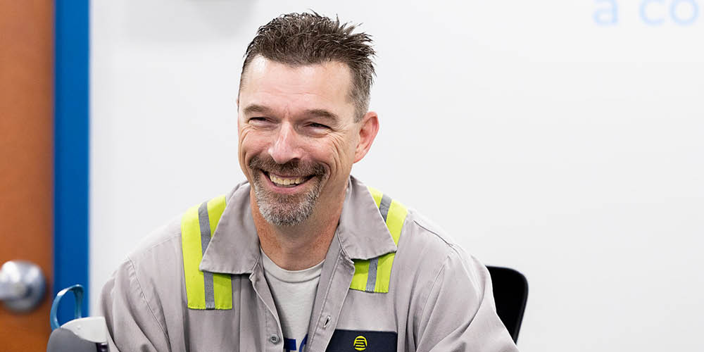 A man with short hair and a goatee, wearing a gray work shirt with yellow reflective stripes, smiles while seated indoors.