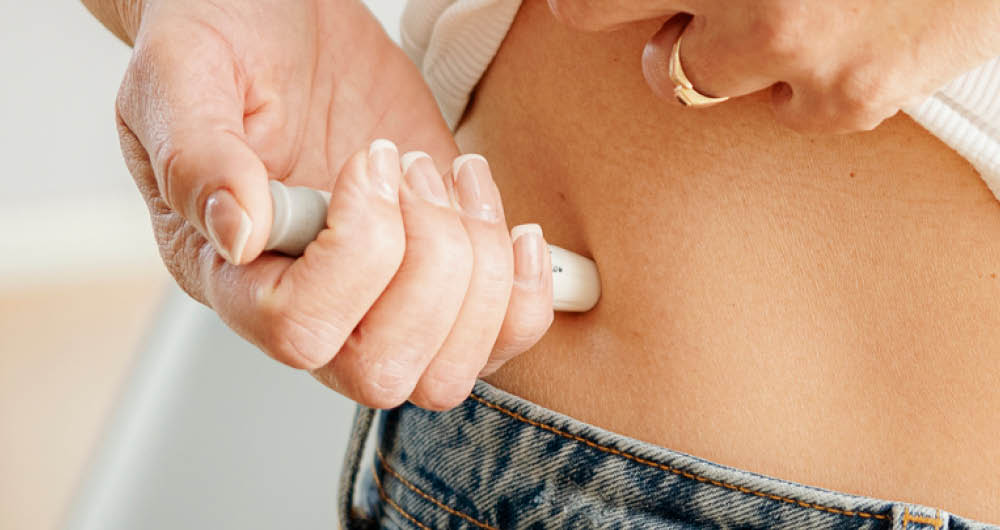 Close-up of a person injecting medication into their abdomen with an insulin pen, wearing jeans and a white top.