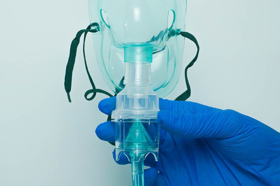 A gloved hand holds a medical oxygen mask with green straps and a transparent valve against a light blue background.