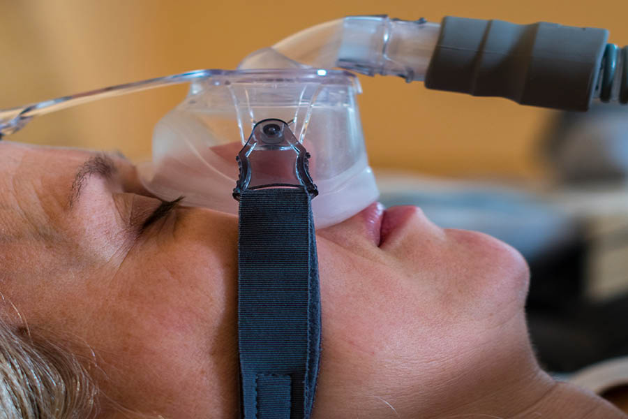 A person lying down wearing a CPAP mask over their nose and mouth, with the mask secured by straps and attached to a hose.