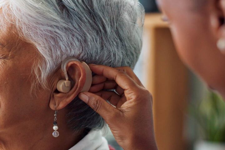 A person fits a behind-the-ear hearing aid on an older adult with short gray hair and a dangling earring.