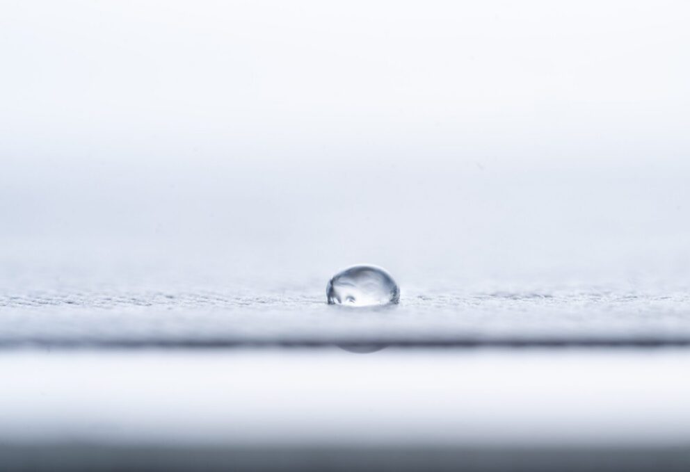 A single water droplet rests on a smooth, light-colored surface with a soft, blurred background.