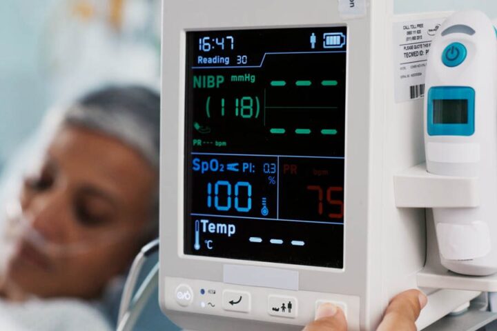 A close-up of a hospital monitor displaying vital signs next to a patient lying in bed wearing an oxygen nasal cannula.