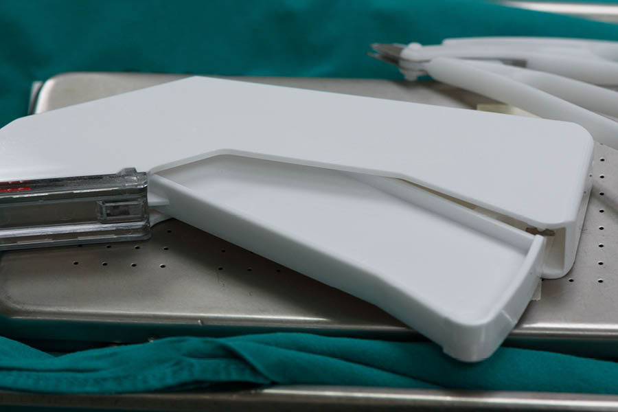 A surgical stapler rests on a metal tray covered with a green surgical drape, with additional white instruments in the background.