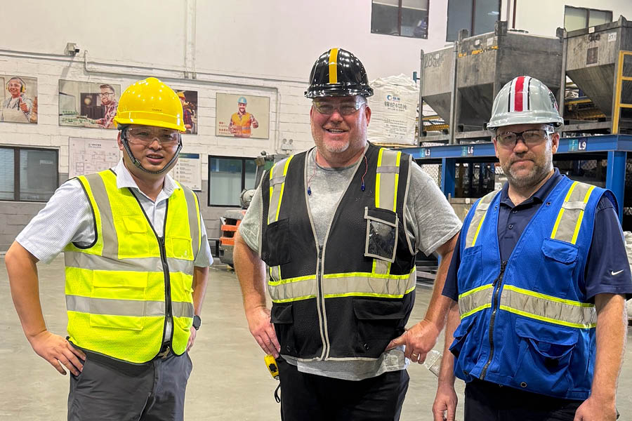 Three men wearing safety vests and helmets stand together in an industrial warehouse setting.