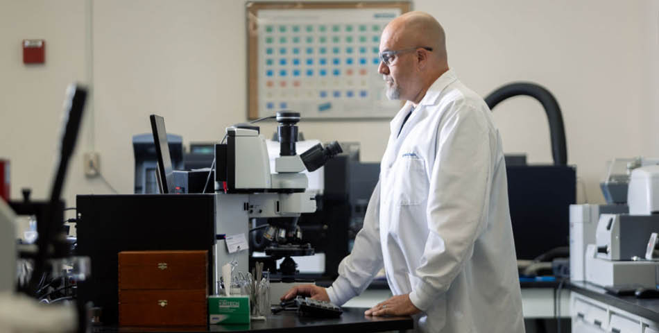 A person in a white lab coat stands at a microscope workstation, typing on a keyboard in a laboratory setting.