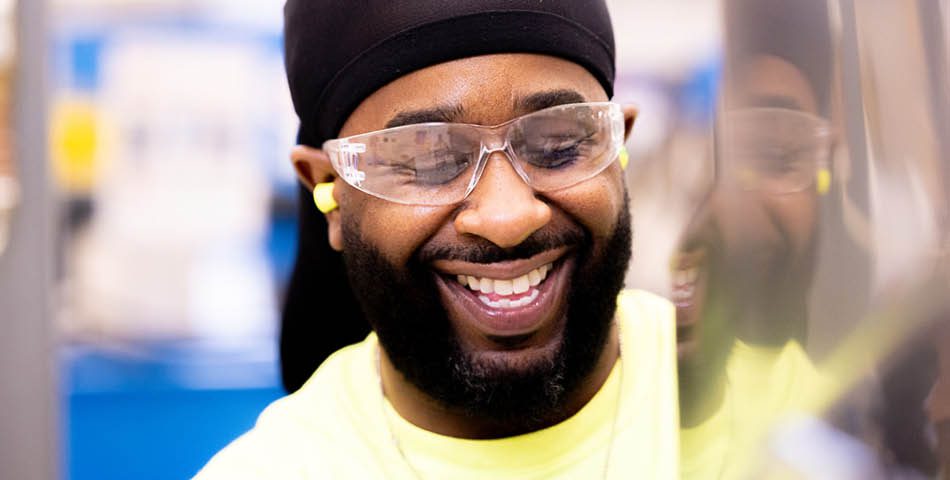 A man wearing safety glasses, a black head covering, and yellow earplugs smiles while working in an industrial setting.