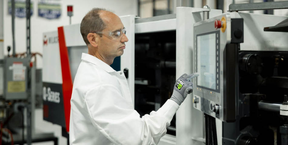 A person wearing safety glasses and gloves operates a touchscreen control panel on industrial machinery in a manufacturing facility.