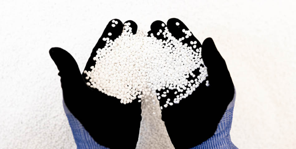 A person wearing black gloves and a blue sleeve holds a pile of white plastic pellets in their cupped hands against a white background.