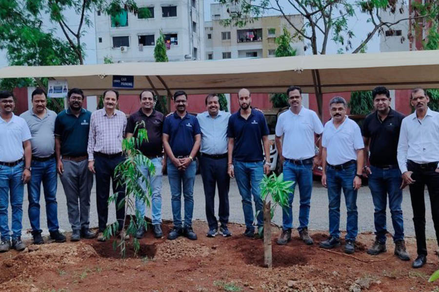 A group of men stand in a row outdoors behind two small, newly planted trees, with buildings and parked cars visible in the background.