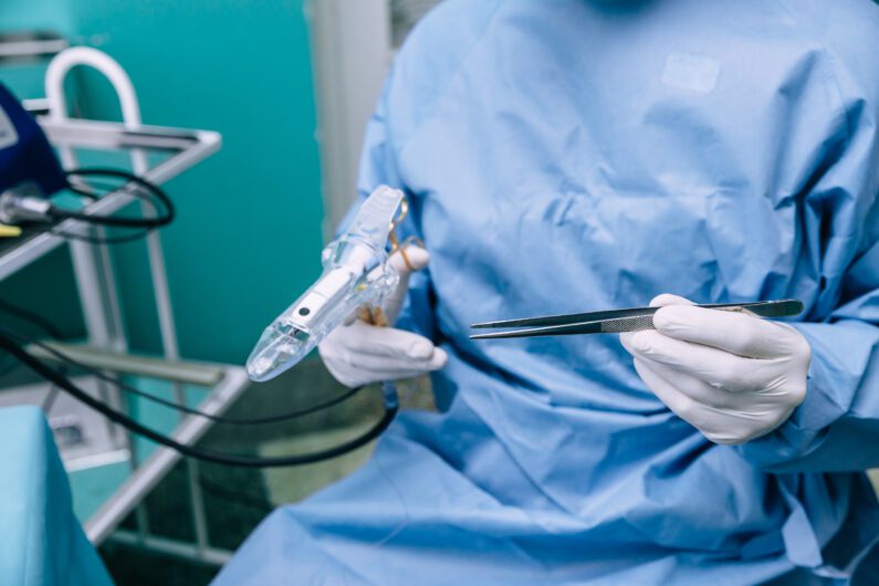 A medical professional in blue scrubs holds a surgical instrument and forceps during a procedure in an operating room.