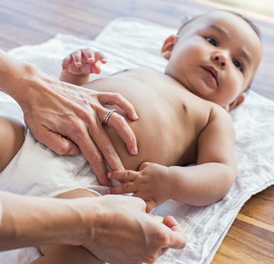 Adult hands changing a baby's diaper while the baby lies on a white cloth on a wooden floor.