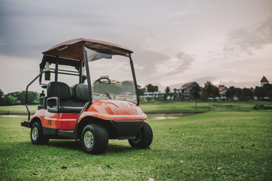 A red golf cart is parked on a grassy golf course with buildings and trees visible in the background under a cloudy sky.