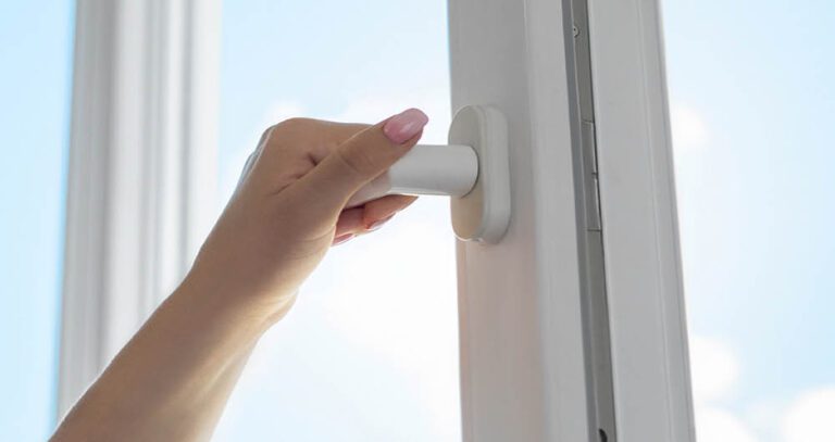 A hand with pink nail polish is turning the handle of a white window to open or close it, with a blue sky visible outside.