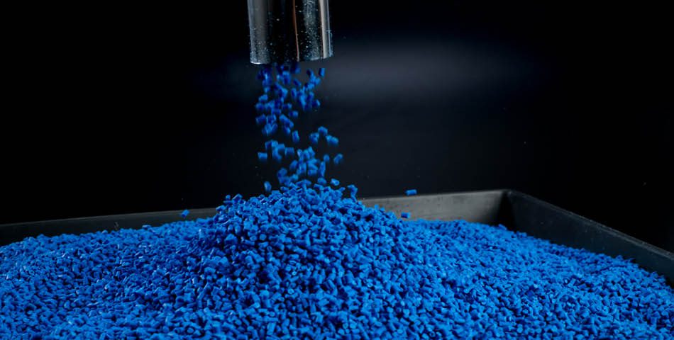 Blue plastic pellets are being dispensed from a metal spout into a pile on a black tray.