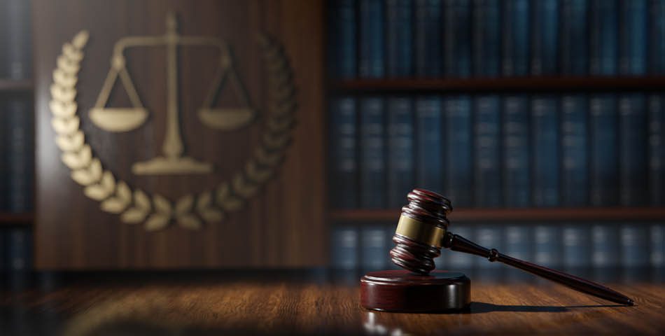 A judge’s gavel rests on a wooden desk with a blurred background of bookshelves and a scales of justice symbol on a wooden panel.