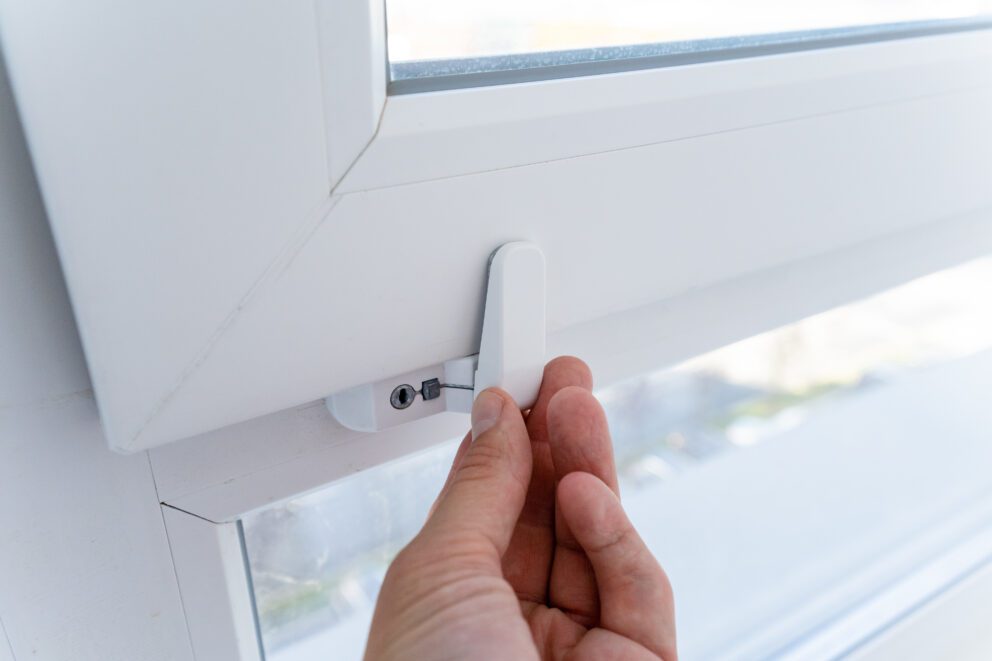 A hand turning the white locking latch of a closed plastic-framed window.