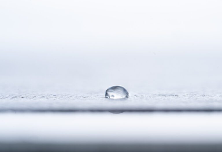A single water droplet rests on a smooth, light-colored surface with a soft, blurred background.