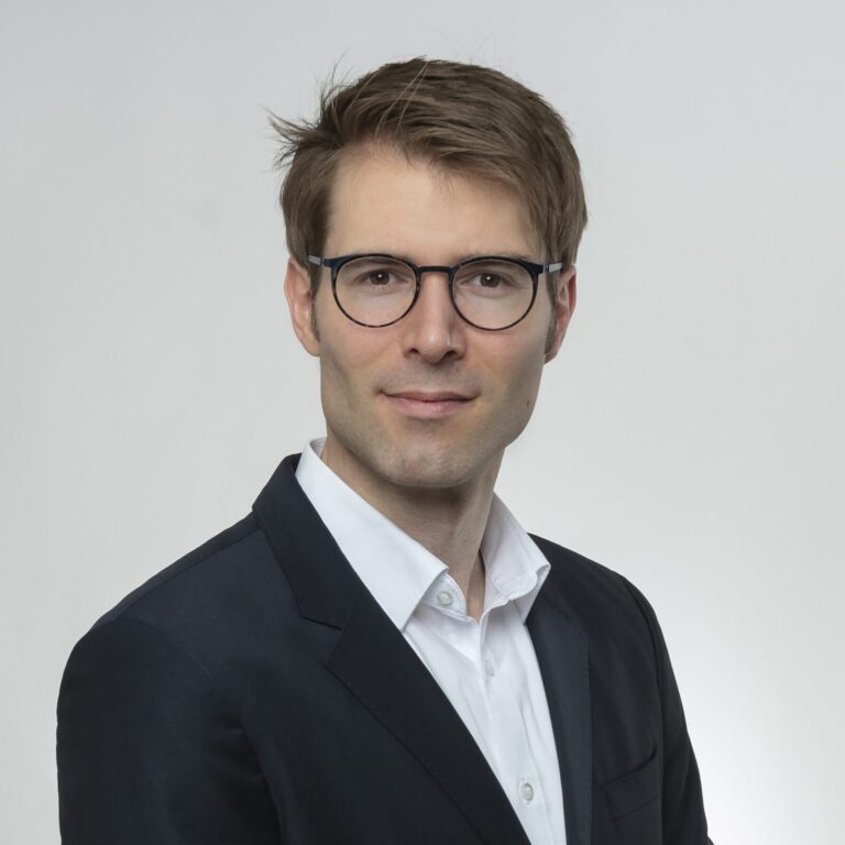 A man with short brown hair and glasses, identified as Kilian Lamprecht, Vice President of Mergers & Acquisitions at Americhem, wears a dark suit jacket and white shirt while posing against a plain light background.